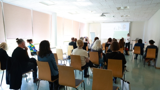 Auditorija seminārā, cilvēki sēž uz krēsliem, skatās uz ekrānu
