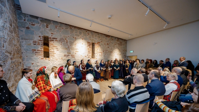 Cilvēki sēž telpā ar mūra sienām - publika un pretī folkloras kopas