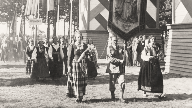 Melnbalta vēsturiska fotogrāfija, kurā cilvēki tautastērpā nes svētku karogu