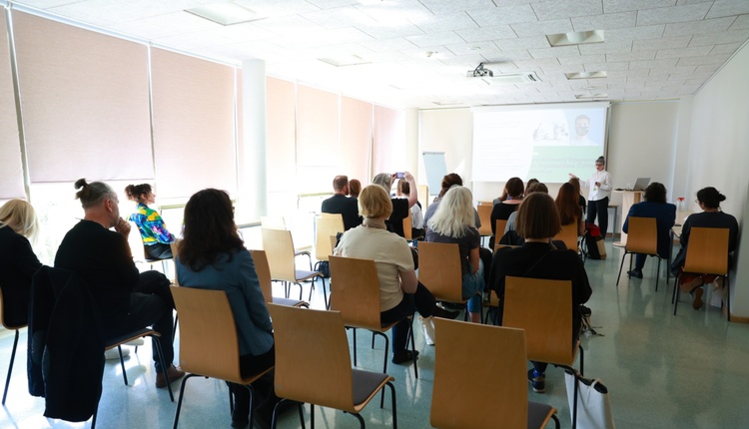 Auditorija seminārā, cilvēki sēž uz krēsliem, skatās uz ekrānu
