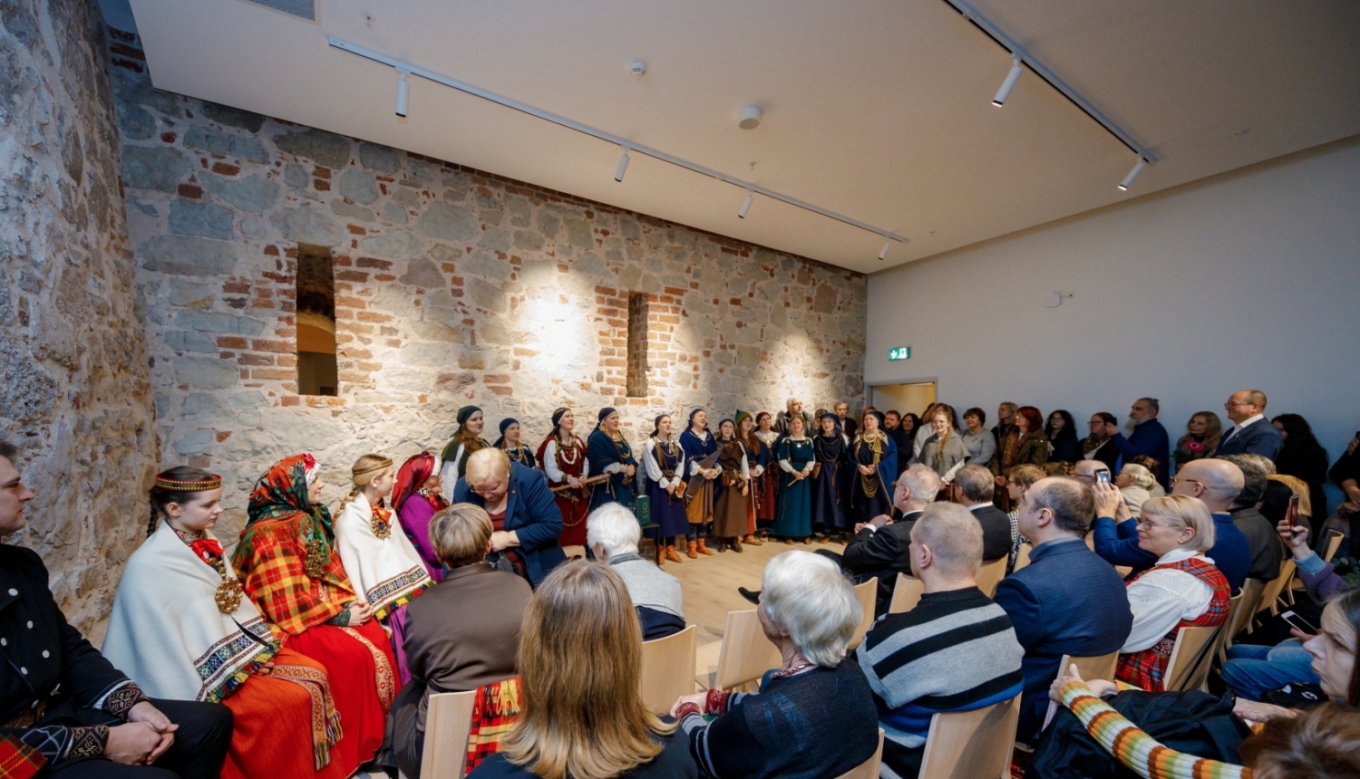 Cilvēki sēž telpā ar mūra sienām - publika un pretī folkloras kopas