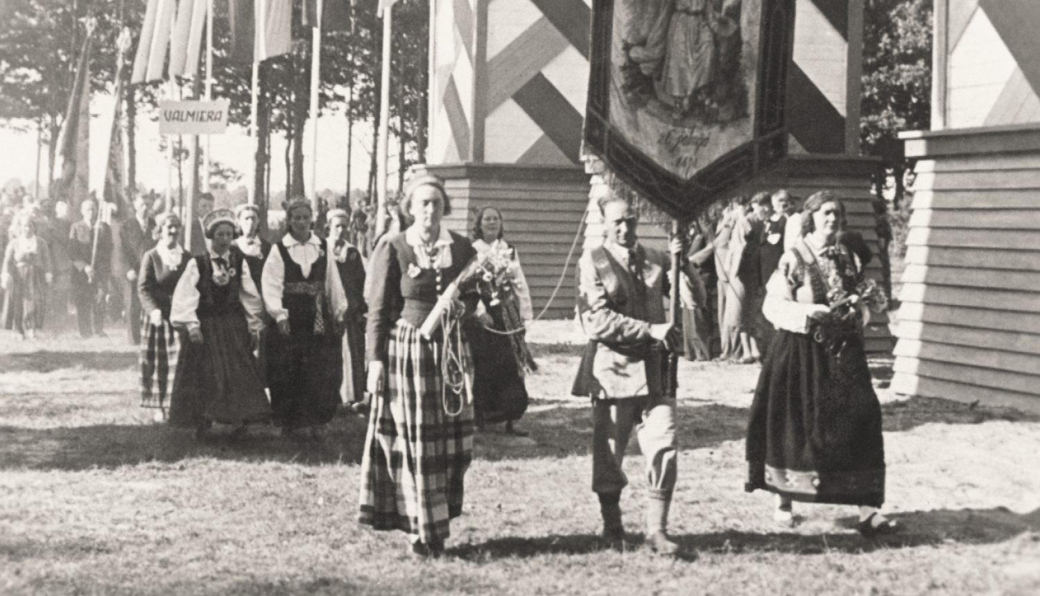 Melnbalta vēsturiska fotogrāfija, kurā cilvēki tautastērpā nes svētku karogu