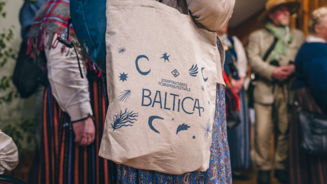 Tuvplānā linu auduma maisiņš ar uzrakstu Baltica. Maisiņš karājas plecā sievietei tautastērpā