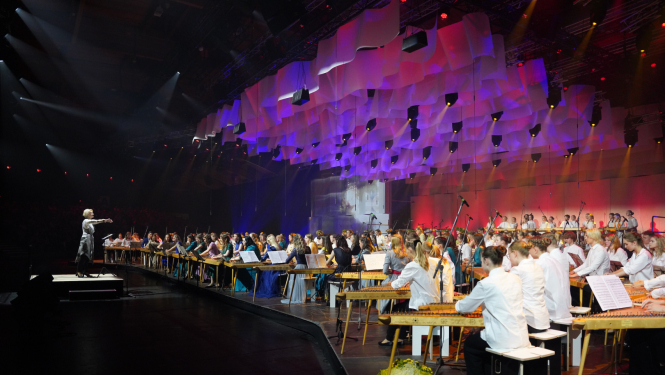 Diriģente diriģē koklētāju kopkors, kas atrodas uz krāšņi izgaismotas skatuves Dziesmu svētkos.