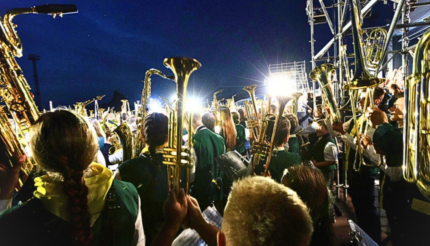Pūtēju orķestru mūziķi tur rokās gaisā paceltus mūzikas instrumentus. Tie  spoži spīd uz tumši zilo debesu fona