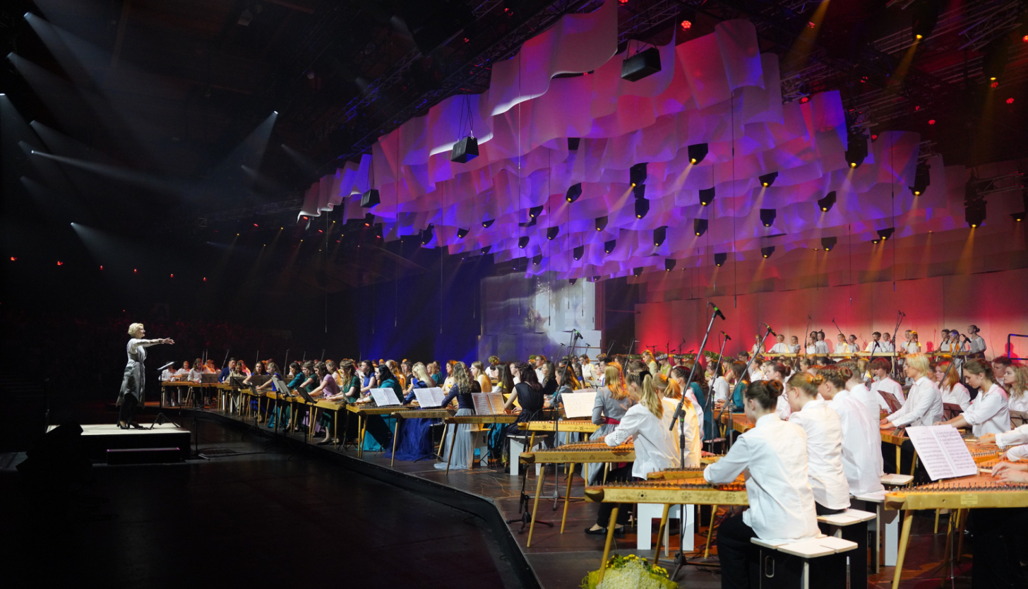 Diriģente diriģē koklētāju kopkors, kas atrodas uz krāšņi izgaismotas skatuves Dziesmu svētkos.